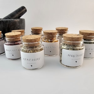 May include: Eight small glass jars with cork lids filled with dried herbs and flowers. The jars are labeled with the names of the herbs and flowers, including Blue Lotus, Rose Petal, Centaury, Calendula, Mugwort, and Alfalfa.