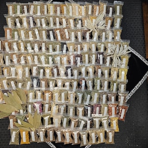 May include: An overhead shot of a collection of small, clear plastic bags filled with various dried herbs and spices. The bags are arranged in a grid pattern, with a few decorative leaves and sprigs. The overall color palette is earthy.