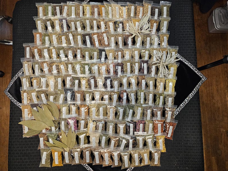 May include: An overhead shot of a collection of small, clear plastic bags filled with various dried herbs and spices. The bags are arranged in a grid pattern, with a few decorative leaves and sprigs. The overall color palette is earthy.