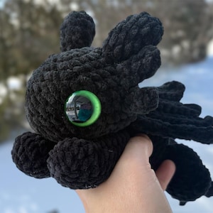 May include: A black crocheted plush dragon toy featuring a large, round head with prominent ear-like appendages and a single, vibrant green plastic eye. The creature has a textured, yarn-based body with small wings and a tail, crafted in a soft style.