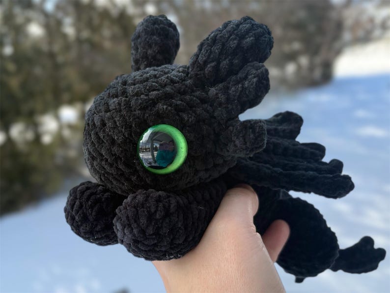 May include: A black crocheted plush dragon toy featuring a large, round head with prominent ear-like appendages and a single, vibrant green plastic eye. The creature has a textured, yarn-based body with small wings and a tail, crafted in a soft style.