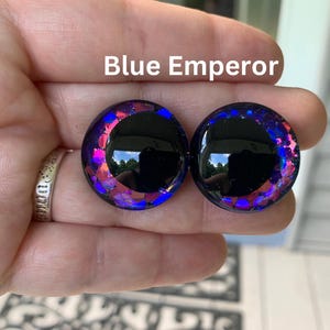 May include: A pair of circular ear plugs labeled Blue Emperor, featuring a smooth black center surrounded by a vibrant border of iridescent blue and purple glitter flakes that catch the light, creating a reflective and multi-dimensional appearance.