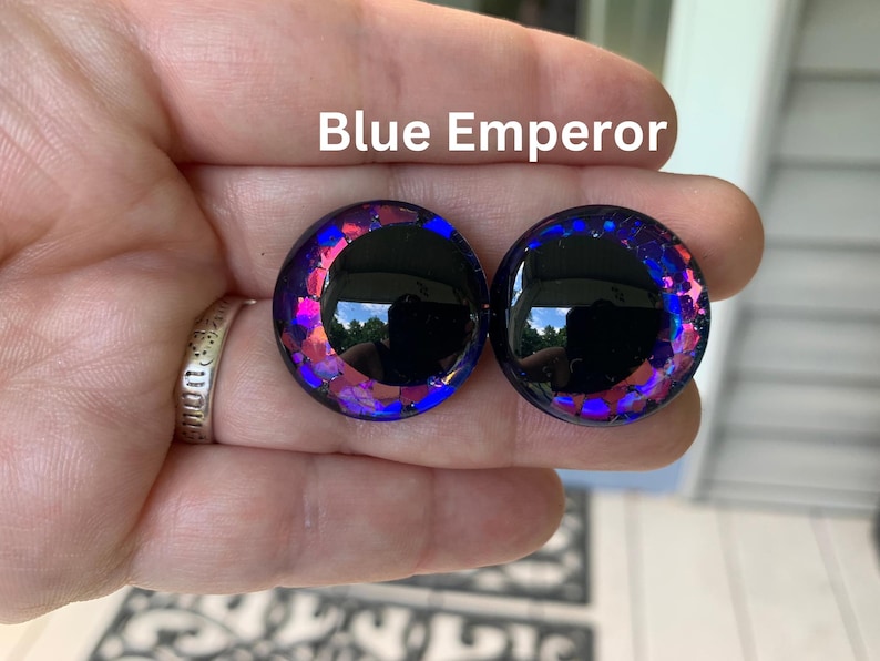 May include: A pair of circular ear plugs labeled Blue Emperor, featuring a smooth black center surrounded by a vibrant border of iridescent blue and purple glitter flakes that catch the light, creating a reflective and multi-dimensional appearance.