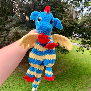 May include: A handmade blue, yellow, and red crochet dragon. The dragon has blue fur with yellow and blue stripes on its body and legs. It has yellow wings, red claws, and a red crest. The dragon is a whimsical, soft toy.