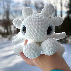 May include: A white crocheted plush toy featuring a rounded head with six protruding branch-like appendages, two large dark blue glossy eyes, and a soft textured yarn surface. The creature has a compact body with small rounded limbs.