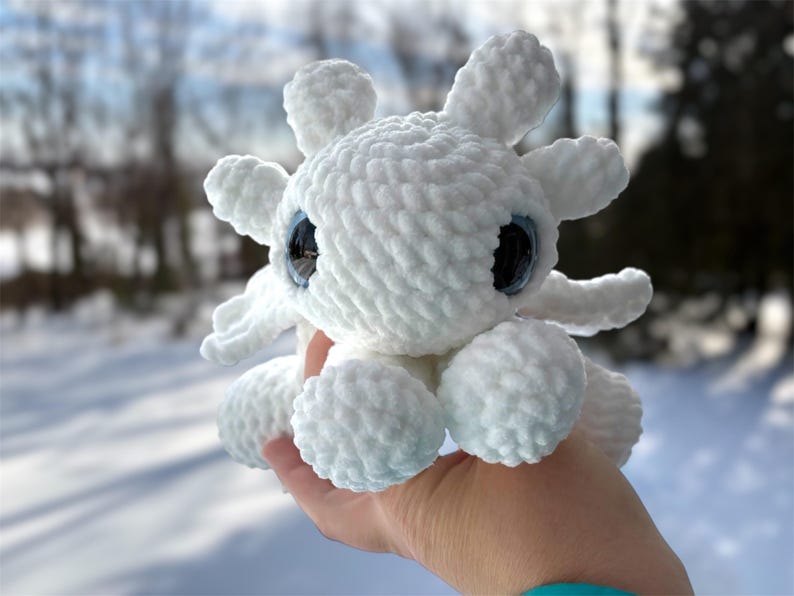 May include: A white crocheted plush toy featuring a rounded head with six protruding branch-like appendages, two large dark blue glossy eyes, and a soft textured yarn surface. The creature has a compact body with small rounded limbs.