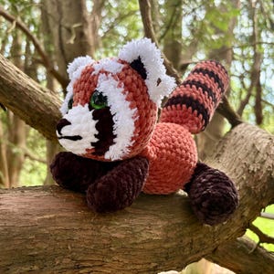 Könnte beinhalten: Ein handgefertigtes gehäkeltes Rotpanda-Plüschtier mit weißen Akzenten, schwarzen Markierungen und grünen Augen. Das Plüschtier sitzt auf einem Ast und zeigt seine detaillierte Verarbeitung und lebendigen Farben. Der Körper des Spielzeugs besteht aus einem weichen, strukturierten Material.
