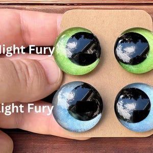 May include: Four circular cabochon eyes mounted on a brown card. The top two are bright green with large black pupils labeled Night Fury. The bottom two are pale blue with large black pupils labeled Light Fury. Each eye has a glossy, reflective finish.