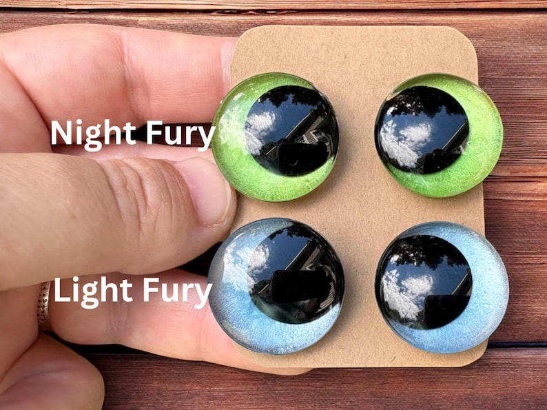 May include: Four circular cabochon eyes mounted on a brown card. The top two are bright green with large black pupils labeled Night Fury. The bottom two are pale blue with large black pupils labeled Light Fury. Each eye has a glossy, reflective finish.