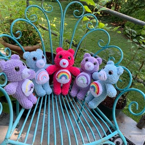 Peut inclure: Cinq ours en peluche au crochet faits à la main dans des tons de rose, de violet et de bleu, chacun avec une application arc-en-ciel sur le ventre. Les ours sont disposés sur une chaise en métal turquoise à l'extérieur, avec de la verdure en arrière-plan.
