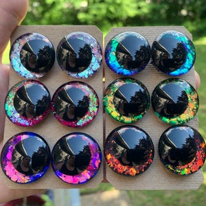 May include: A set of twelve circular cabochons arranged on two cardboard sheets. Each piece features a large black pupil surrounded by a colorful iris filled with iridescent, reflective glitter flakes in shades of pink, purple, blue, green, and orange.
