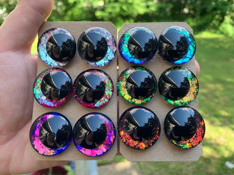 May include: A set of twelve circular cabochons arranged on two cardboard sheets. Each piece features a large black pupil surrounded by a colorful iris filled with iridescent, reflective glitter flakes in shades of pink, purple, blue, green, and orange.