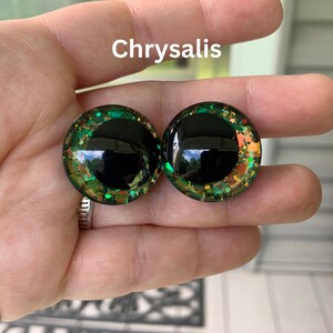 May include: A pair of round, domed cabochons labeled Chrysalis, featuring a large, solid black circular pupil centered within a vibrant iris composed of green, gold, and copper-toned glitter flakes embedded in a clear, glossy resin material.
