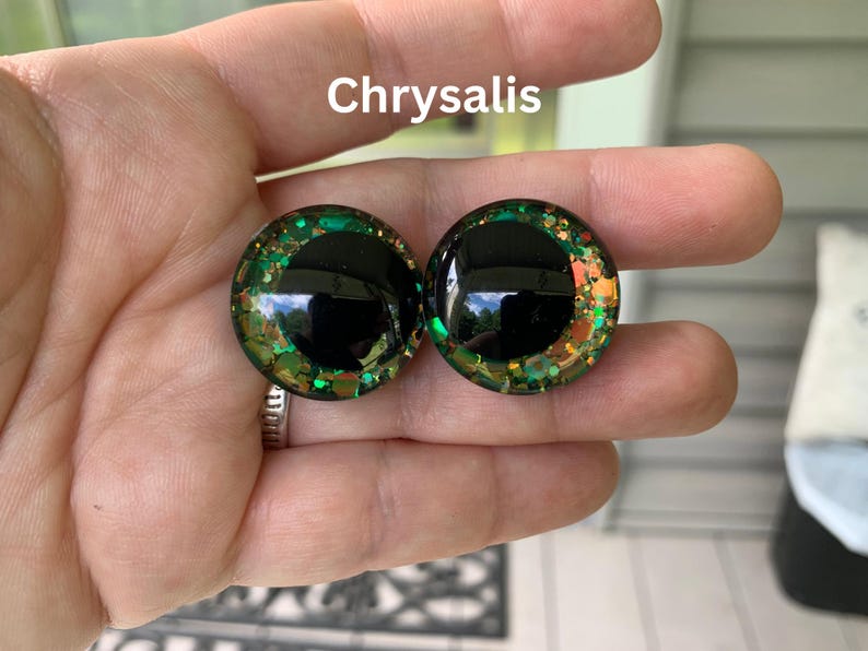 May include: A pair of round, domed cabochons labeled Chrysalis, featuring a large, solid black circular pupil centered within a vibrant iris composed of green, gold, and copper-toned glitter flakes embedded in a clear, glossy resin material.