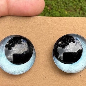 May include: A pair of circular stud earrings featuring a glossy black dome center surrounded by a shimmering light blue metallic border. The earrings are mounted on a rectangular piece of brown cardboard for display and storage purposes.