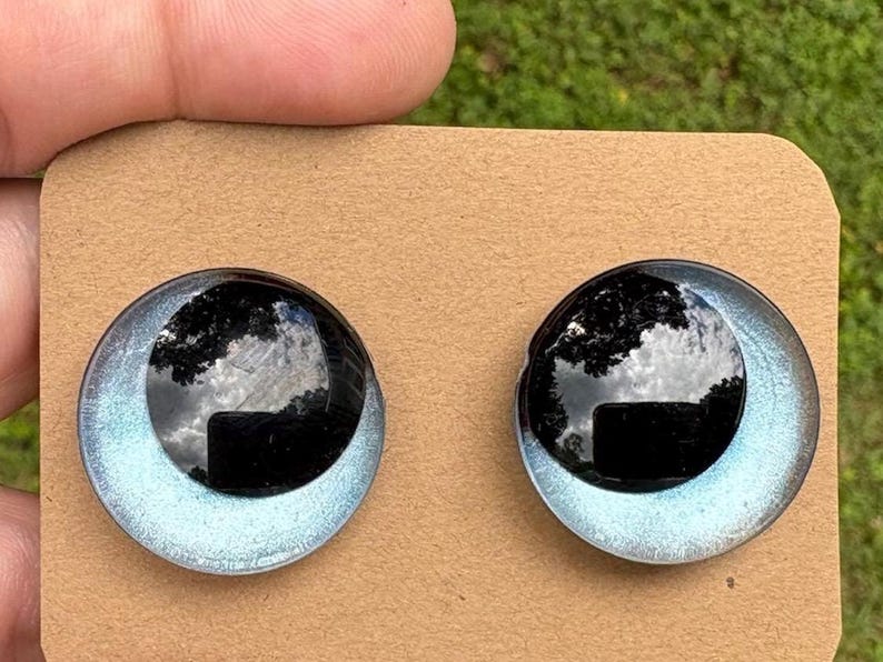 May include: A pair of circular stud earrings featuring a glossy black dome center surrounded by a shimmering light blue metallic border. The earrings are mounted on a rectangular piece of brown cardboard for display and storage purposes.