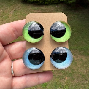 May include: Four circular cabochons arranged on a rectangular tan card. The top two cabochons feature a bright lime green iris with a large black pupil. The bottom two cabochons feature a light blue iris with a large black pupil. Each piece has a smooth, glossy, domed surface.