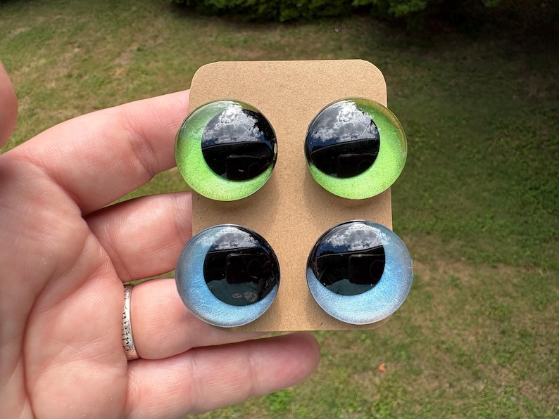 May include: Four circular cabochons arranged on a rectangular tan card. The top two cabochons feature a bright lime green iris with a large black pupil. The bottom two cabochons feature a light blue iris with a large black pupil. Each piece has a smooth, glossy, domed surface.