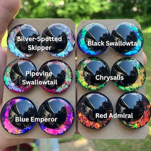 May include: Twelve circular cabochons arranged in pairs on two cardboard cards. Each piece features a black pupil and a colorful, glittery iris. Labels identify them as Silver-Spotted Skipper, Black Swallowtail, Pipevine Swallowtail, Chrysalis, Blue Emperor, and Red Admiral.