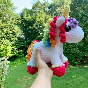 Puede incluir: Un unicornio blanco tejido a ganchillo con una melena, cola y cascos de arcoíris. El unicornio tiene un cuerno amarillo y ojos grandes con pupilas negras. El juguete se sostiene en una mano sobre un fondo de árboles y hierba verdes.