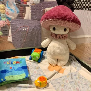 May include: A crocheted mushroom toy with a pink and white cap and a white body. The toy is sitting on a white and blue blanket with a toy truck and a yellow toy animal.