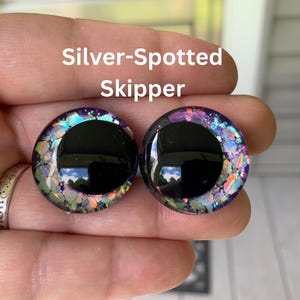 May include: Two circular cabochons labeled Silver-Spotted Skipper, featuring a large solid black pupil surrounded by a translucent iris filled with iridescent, multi-colored glitter flakes that shimmer in shades of purple, blue, green, and silver.