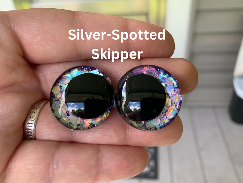May include: Two circular cabochons labeled Silver-Spotted Skipper, featuring a large solid black pupil surrounded by a translucent iris filled with iridescent, multi-colored glitter flakes that shimmer in shades of purple, blue, green, and silver.