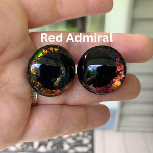 May include: Two circular black plugs with a glossy finish, featuring embedded multi-colored glitter accents in shades of red, orange, and green along the edges. The text Red Admiral is printed in white above the circular jewelry pieces.