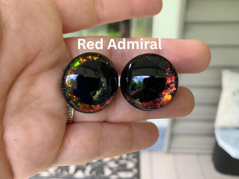May include: Two circular black plugs with a glossy finish, featuring embedded multi-colored glitter accents in shades of red, orange, and green along the edges. The text Red Admiral is printed in white above the circular jewelry pieces.