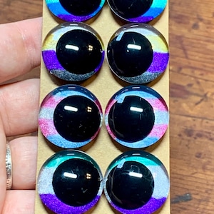 Puede incluir: Ocho cabujones de vidrio con pupila negra e iris de colores. Los iris presentan un arcoíris de colores, incluyendo rojo, naranja, amarillo, verde, azul y morado. Los cabujones están dispuestos en dos filas de cuatro sobre un soporte de cartón.