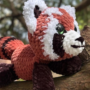 May include: A handmade crochet red panda plush toy with white, brown, and black accents. The plush animal has green eyes and is lying on a tree branch. The plush toy is made of soft, textured yarn.