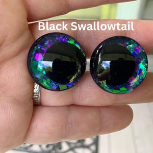 May include: Two circular cabochons labeled Black Swallowtail, featuring a large solid black center surrounded by a ring of iridescent purple, blue, and green glitter flakes embedded in a clear, glossy, dome-shaped resin material.