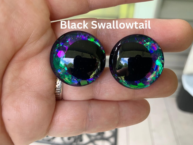 May include: Two circular cabochons labeled Black Swallowtail, featuring a large solid black center surrounded by a ring of iridescent purple, blue, and green glitter flakes embedded in a clear, glossy, dome-shaped resin material.