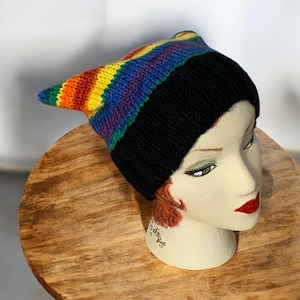 May include: A rainbow striped knit beanie with a black brim and a pointed top. The beanie is on a mannequin head.