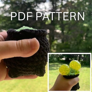 May include: A black crocheted cauldron with green filling, held in a hand. The text "PDF PATTERN" is displayed above the cauldron. A smaller image shows a similar cauldron with yellow orbs.