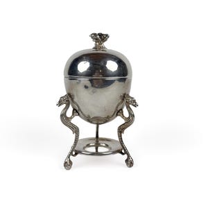Op de afbeelding: Een zilverkleurige, eivormige houder met deksel, ondersteund door een driepootstandaard met drie gestileerde vogelachtige poten. De houder heeft een glad, reflecterend oppervlak en een kleine decoratieve knop bovenop het deksel.