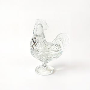 May include: A clear glass rooster-shaped candy dish. The dish features a detailed design with a textured body, a comb, and a tail. It is elevated on a pedestal base, creating a decorative and functional piece for serving or display.