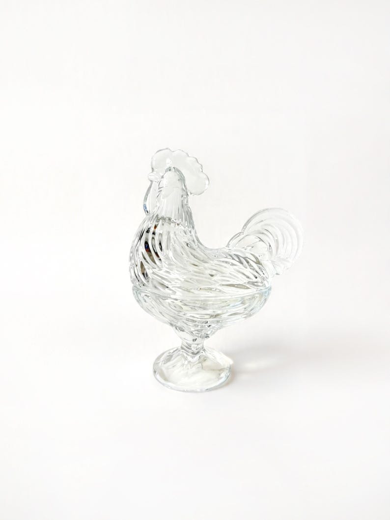 May include: A clear glass rooster-shaped candy dish. The dish features a detailed design with a textured body, a comb, and a tail. It is elevated on a pedestal base, creating a decorative and functional piece for serving or display.