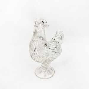 May include: A clear glass rooster figurine with a textured, detailed design. The rooster stands on a pedestal base, showcasing its comb, wattle, and feathered body. The figurine is a decorative item.