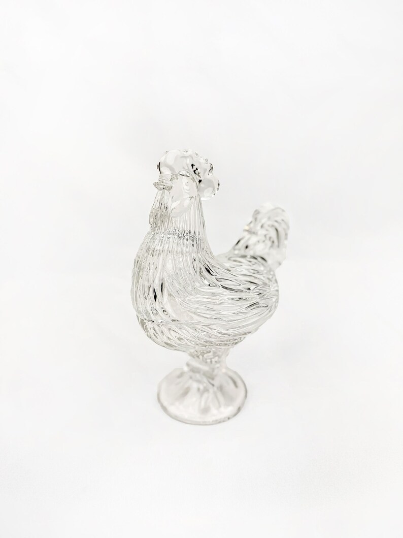 May include: A clear glass rooster figurine with a textured, detailed design. The rooster stands on a pedestal base, showcasing its comb, wattle, and feathered body. The figurine is a decorative item.