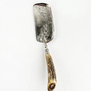 May include: Silver-plated cake server with an ornate floral and scrollwork design on the blade. The handle is made of a light brown antler material. A decorative serving utensil for cakes and pastries.