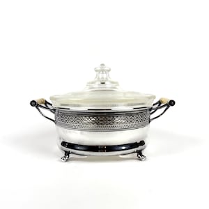 May include: A silver-plated serving dish with a clear glass lid, featuring a decorative filigree band around the upper rim, two side handles with light-coloured accents, and four small feet. The lid has a central knob handle.