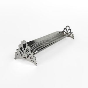 Può includere: Un supporto in metallo color argento con una base lunga, stretta e ondulata per contenere oggetti. Il supporto è sostenuto a ciascuna estremità da una base triangolare decorativa con un intricato motivo a filigrana traforata.