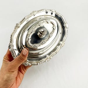 Puede incluir: Una fuente ovalada plateada con tapa. La fuente presenta detalles ornamentados en relieve alrededor de los bordes y en la tapa. Un pomo con forma de cristal adorna el centro de la tapa. La fuente es sostenida por una mano.