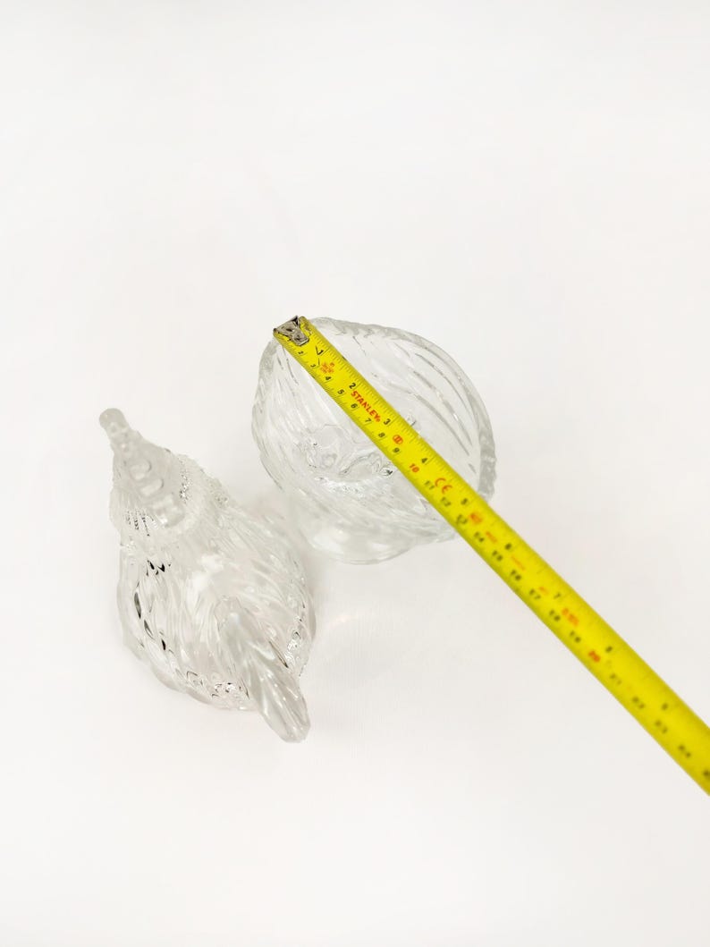 May include: Two clear glass decorative pieces. One is a small bowl with a textured, ridged design. The other is a bird-shaped piece with a textured surface. A yellow tape measure is in the frame, showing measurements in inches.