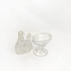 May include: Clear glass hen-shaped dish and a small pedestal bowl. The hen dish has a textured, feathered design. The bowl has a twisted pattern. Both are likely vintage and suitable for serving or display.