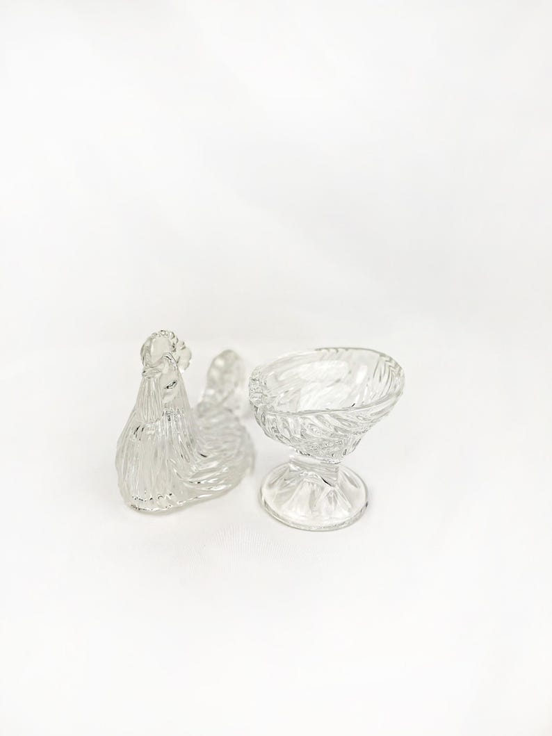 May include: Clear glass hen-shaped dish and a small pedestal bowl. The hen dish has a textured, feathered design. The bowl has a twisted pattern. Both are likely vintage and suitable for serving or display.