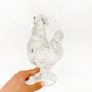 May include: A clear glass hen-shaped decorative object. The hen has a textured, ribbed design and is standing on a pedestal base. The object is transparent and reflects light, creating a sparkling effect. The hen is a decorative item.
