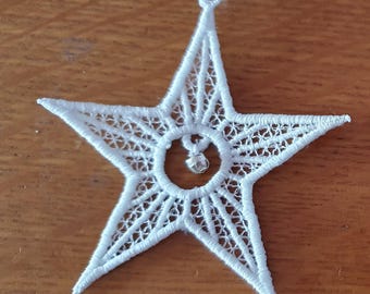 Può includere: Due ornamenti bianchi a forma di stella con intricati dettagli in pizzo. Ogni stella presenta un design circolare centrale con una piccola perla trasparente e un nastro bianco per appenderla. Su una superficie di legno.