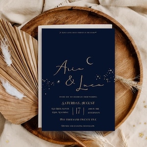 May include: A navy blue wedding invitation with gold script, featuring the names "Avia & Luca" and event details. The invitation is set on a wooden tray with dried palm leaves and feathers, creating a rustic, celestial theme.